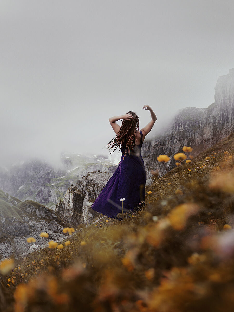 Fine Art Photography by Anna Heimkreiter. The photo shows an ethereal scene of a woman in a flower meadow among giant mountains.